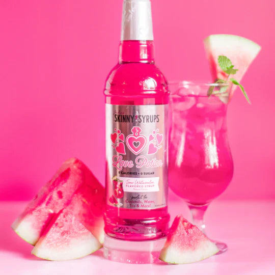 bottle of Sour Love Potion skinny syrup set next to a daiquiri glass and watermelon slices arranged on a a pink background.