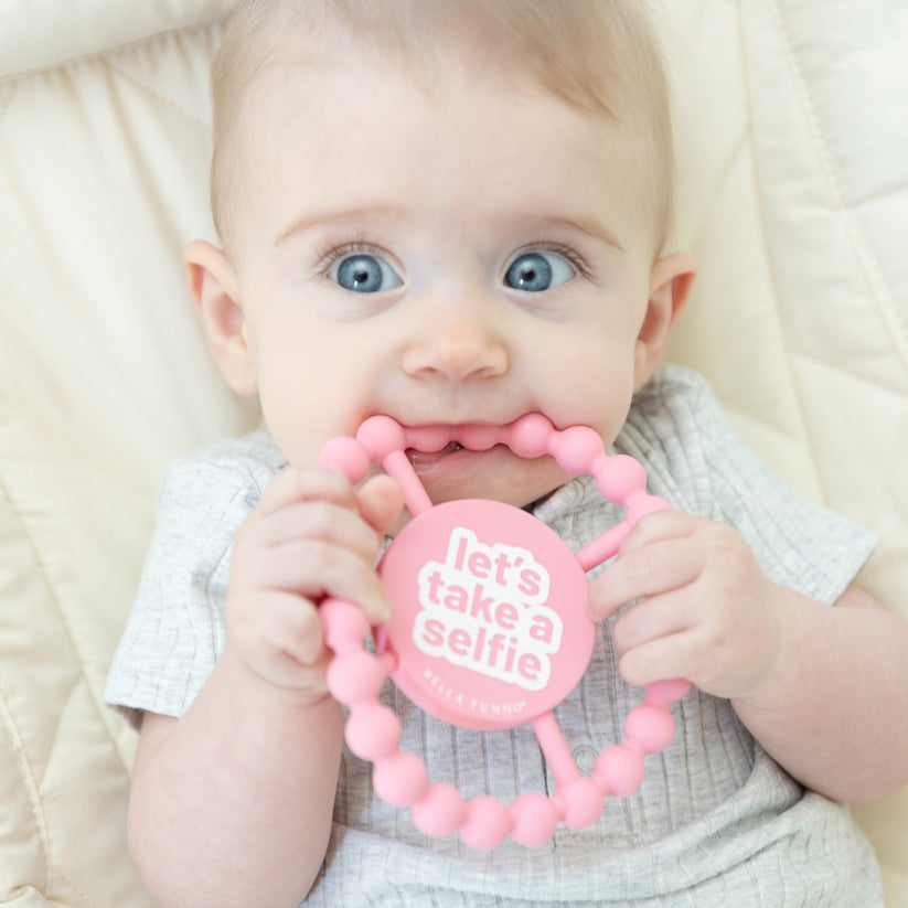 baby laying down chewing on "selfie" teether.