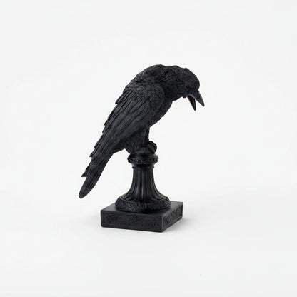 A black crow figurine perched on a decorative pedestal base, shown against a seamless white background.