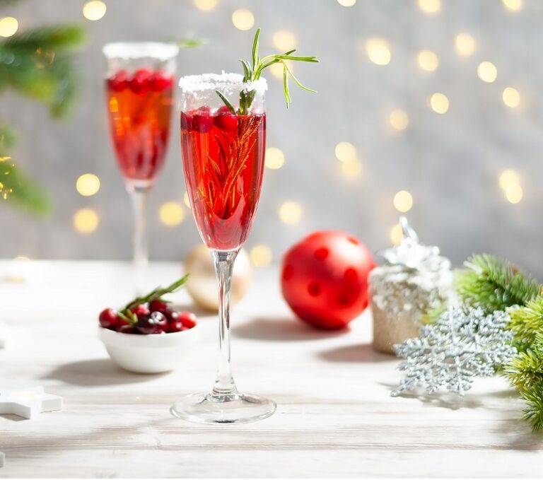 2 glasses of cranberry prosecco set on a table with a bowl of cranberries, Christmas ornaments, and greenery.