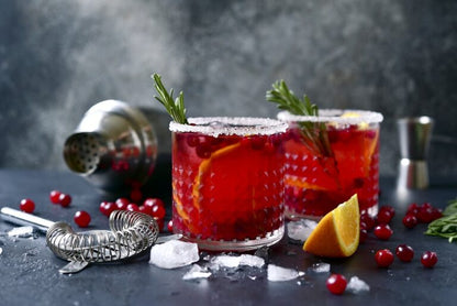 2 glasses filled with cranberry sangria set on a grey counters with cranberries, orange slices, and bar tools scattered around.
