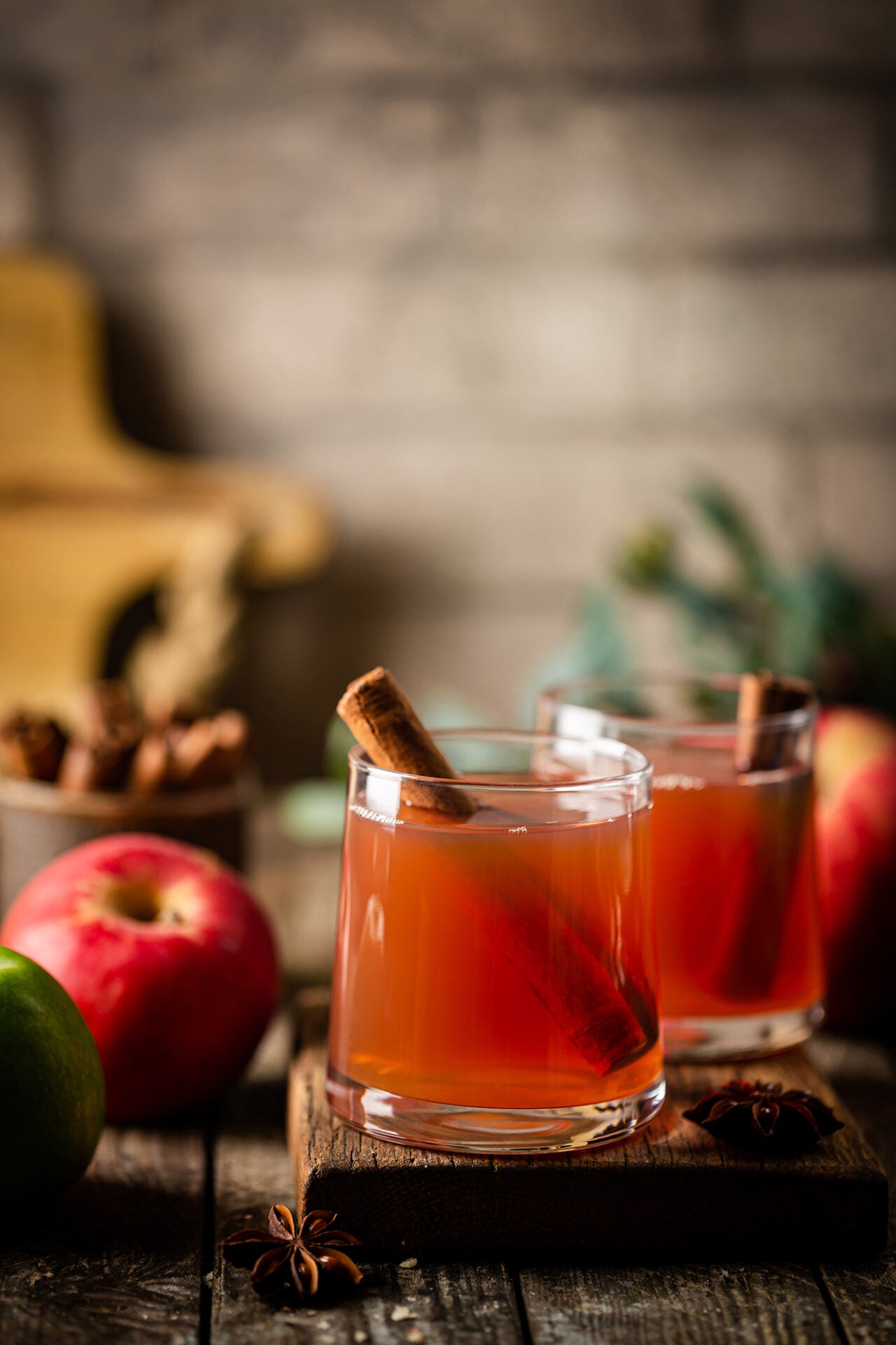 two caramel apple toddy drinks with cinnamon sticks displayed on a wood serving board next to apples