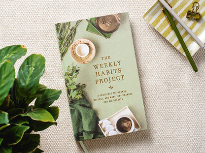 The Weekly Habits Project book laying on a table with pens and a plant