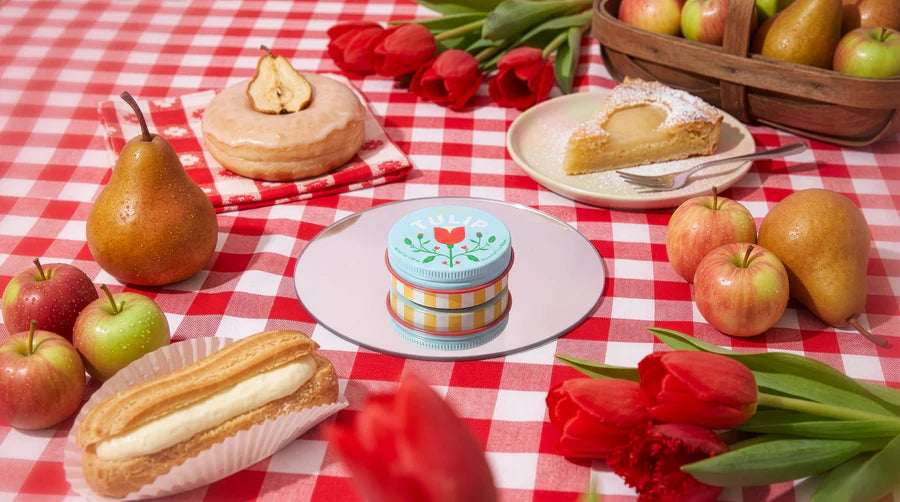 Tulip lip balm set on a red gingham picnic blanket surrounded by apples and pastries.