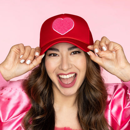 person smiling and wearing red trucker hat with pink heart on it.