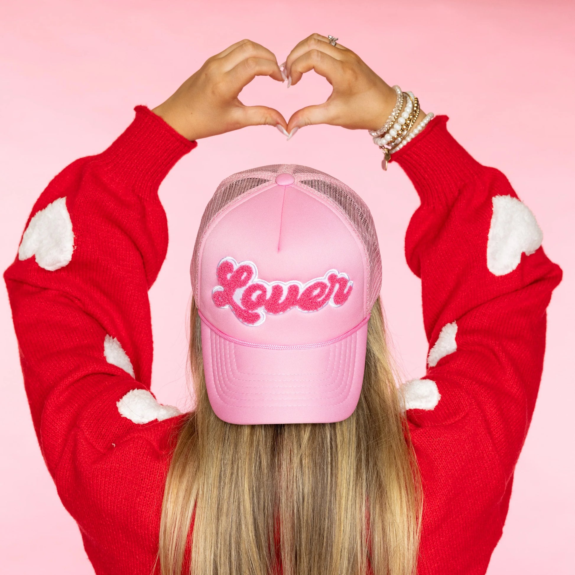 person wearing pink "lover" hat and making heart hands above their head.