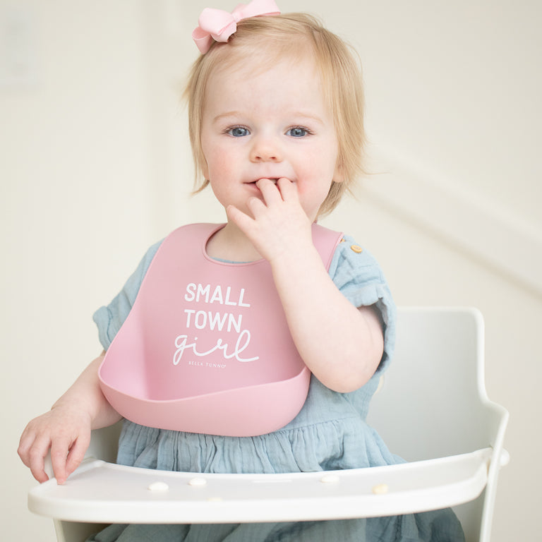 a little girl sitting in a highchair wearing the pink silicone small town girl wonder bib