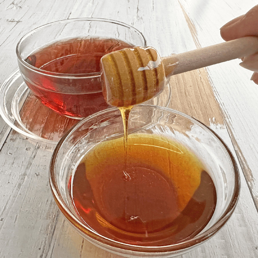 Honey being drizzled from a wooden honey dipper into a glass bowl on a light wooden surface.