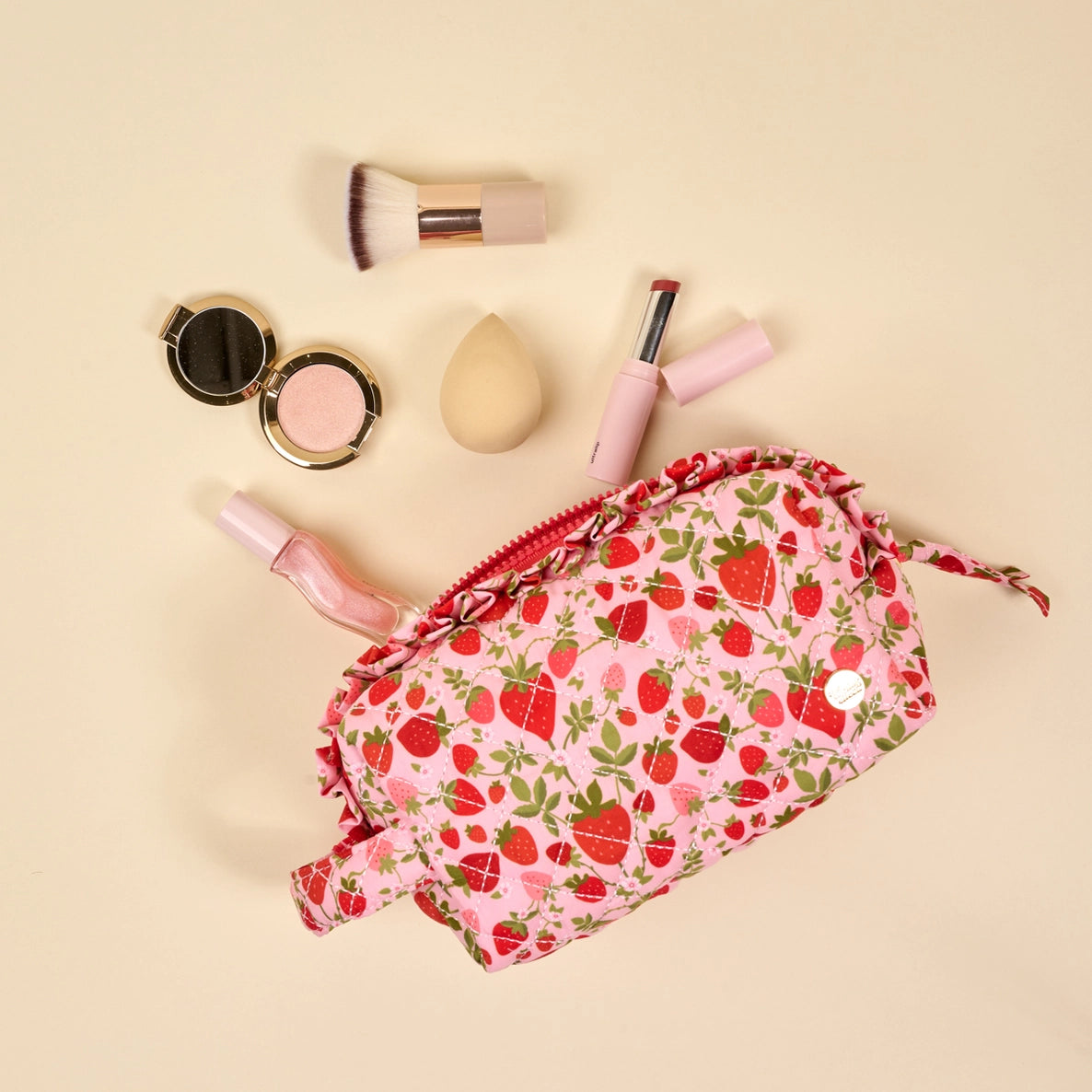 Makeup bag with strawberry pattern next to makeup items on a beige background