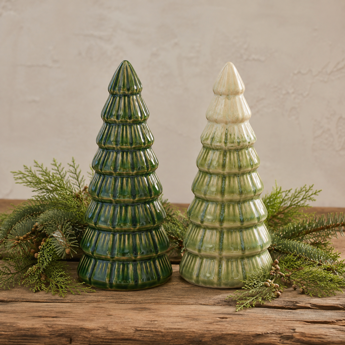 Two ceramic Christmas trees on a rustic wooden surface, arranged with green pine branches. One tree is glazed in dark green, and the other in light green with a cream-colored pointed top.