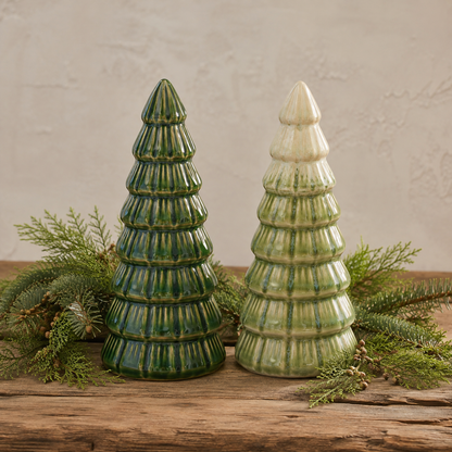 Two ceramic Christmas trees on a rustic wooden surface, arranged with green pine branches. One tree is glazed in dark green, and the other in light green with a cream-colored pointed top.