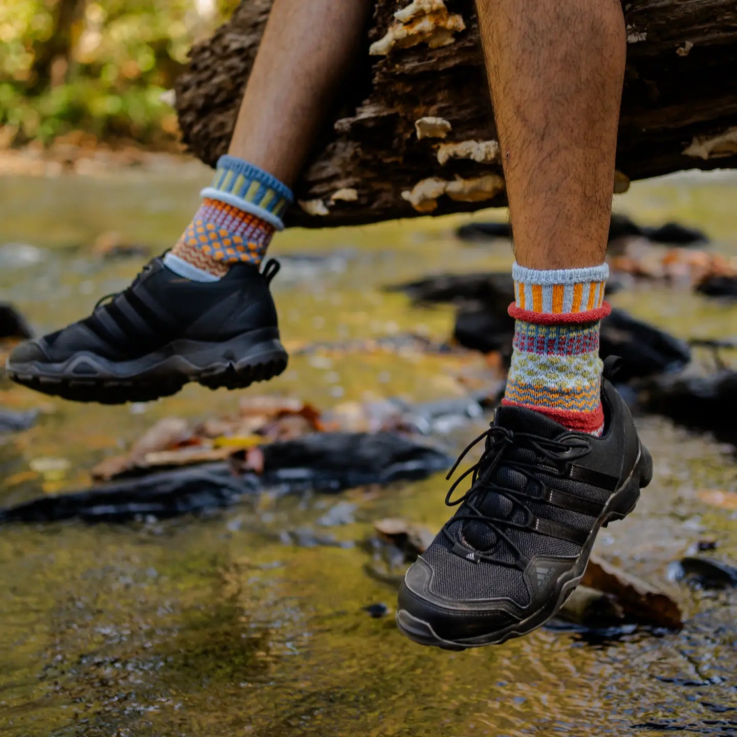 person wearing black athletic shoes dangling the feet off a log over a river