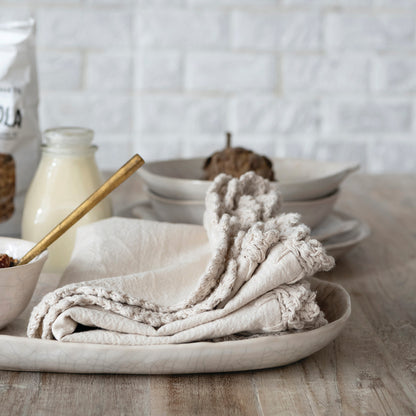 cream colored platter with folded kitchen linens set on it shown on a table with a stack of dishes and milk jug in the background