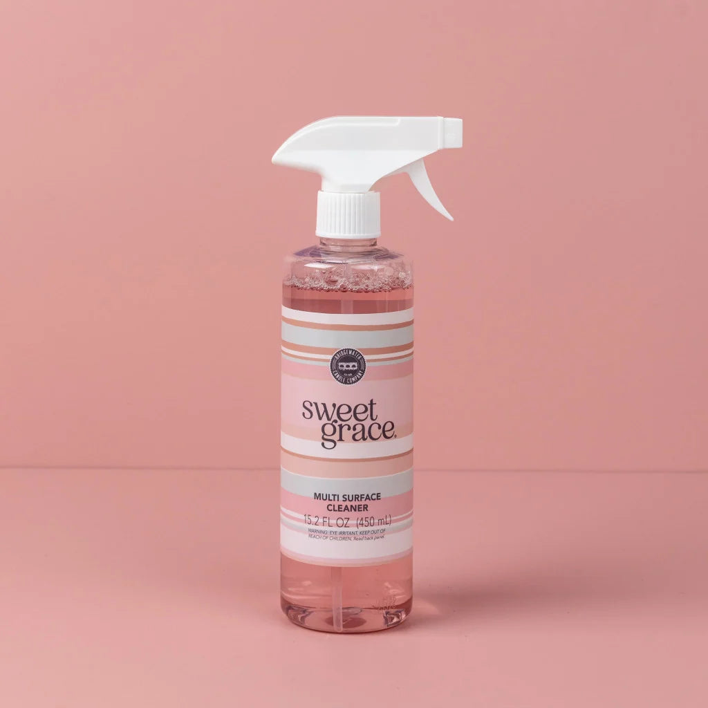 A bottle of Sweet Grace Multi-Surface Cleaner with pink liquid, a white spray trigger top, and a pink and white striped label, set against a solid pink background.