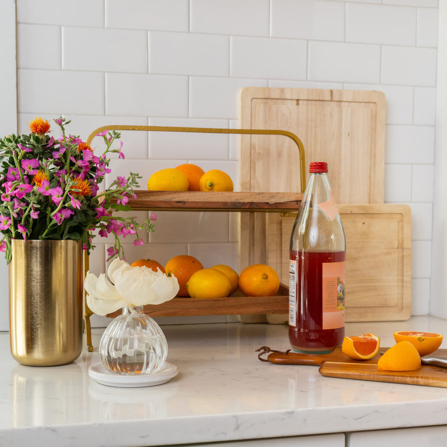 Orange & Honey Flower Diffuser set on a kitchen counter surrounded by flowers, a small shelf filled woth fresh oranges, a cutting board with orange wedges on it, and a bottle of juice.