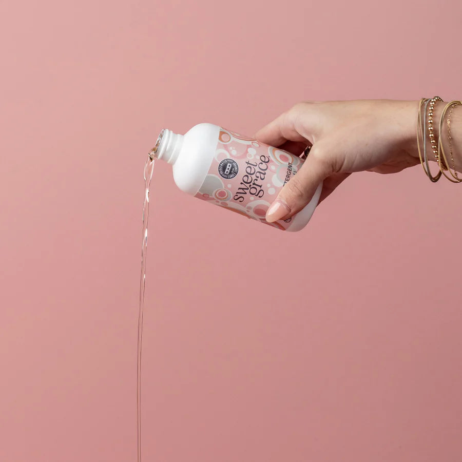 Hand pouring a sweet grace laundry detergent from a bottle against a pink background