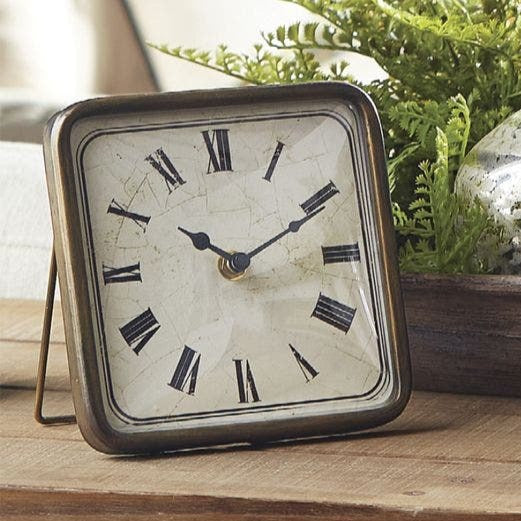 square clock set on a wooden table with greenery next to it.