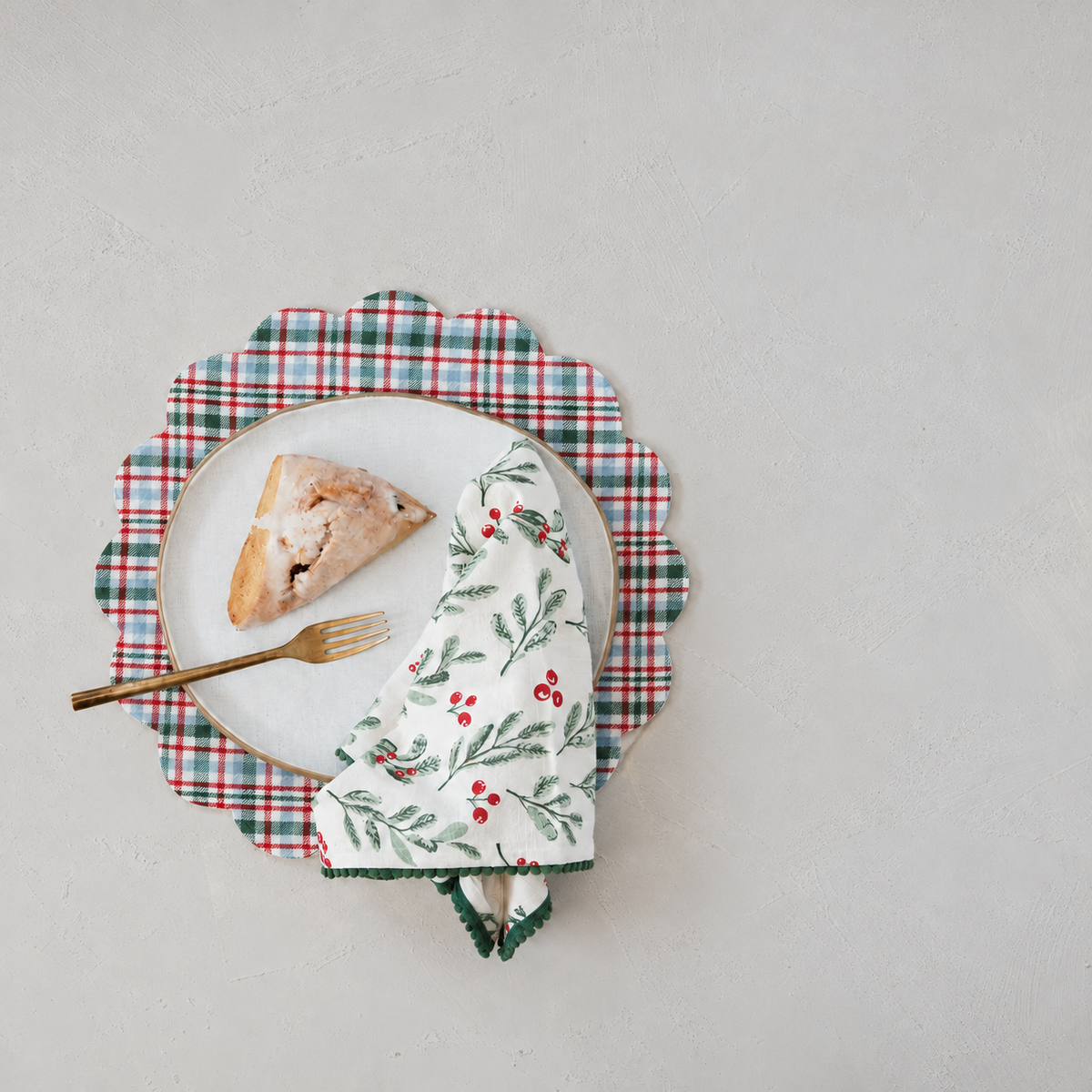 A styled flat-lay featuring a white ceramic plate with a scone, gold-toned fork, and a white napkin with a green and red holly pattern, all arranged on top of a red, green, and white plaid placemat with scalloped edge against a light gray background.