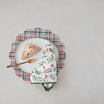 A styled flat-lay featuring a white ceramic plate with a scone, gold-toned fork, and a white napkin with a green and red holly pattern, all arranged on top of a red, green, and white plaid placemat with scalloped edge against a light gray background.