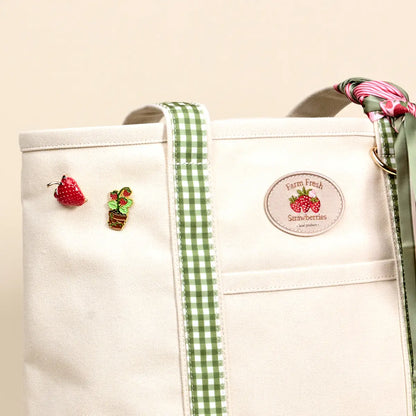 close-up of Off-White tote bag with green gingham strap, strawberry-themed decorations, and a label on a beige background.