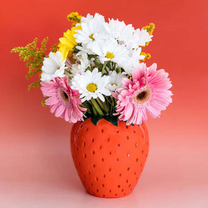 A ceramic vase designed to resemble a strawberry, with a variety of flowers arranged in it, placed against a red background.