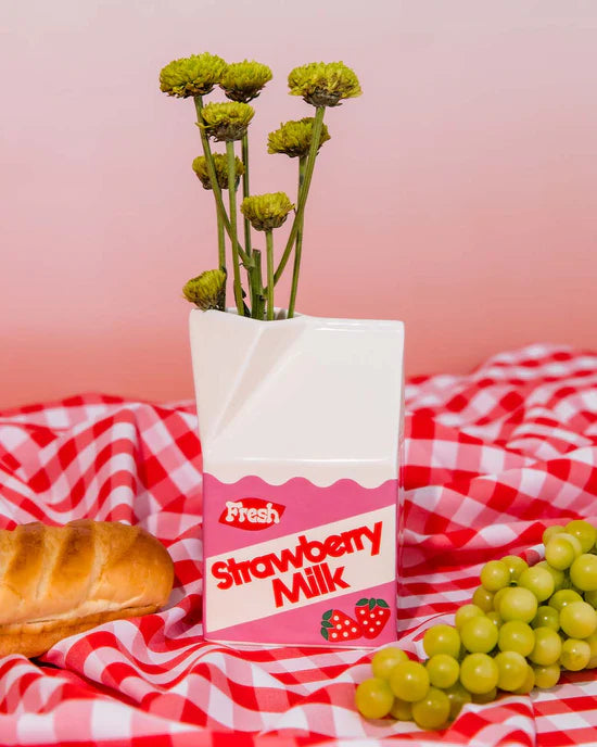 white carton shaped vase with pink and red design and "fresh strawberry milk" painter on it and filled with small flowers arranged on a red check cloth with bread and grapes.