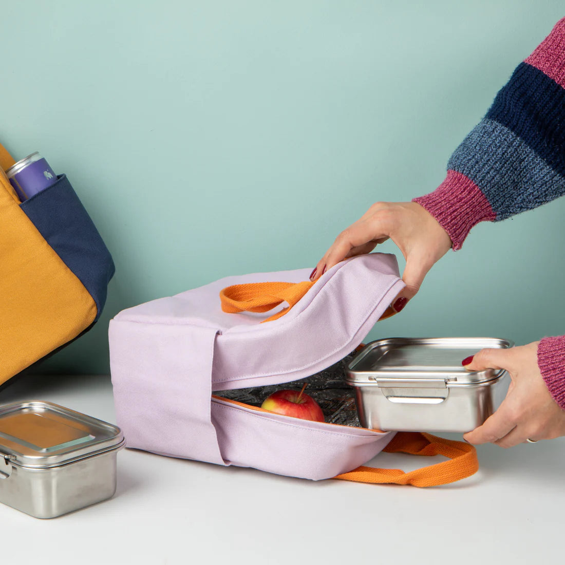 Person opening a lavender lunch bag with containers and food items on a light green background