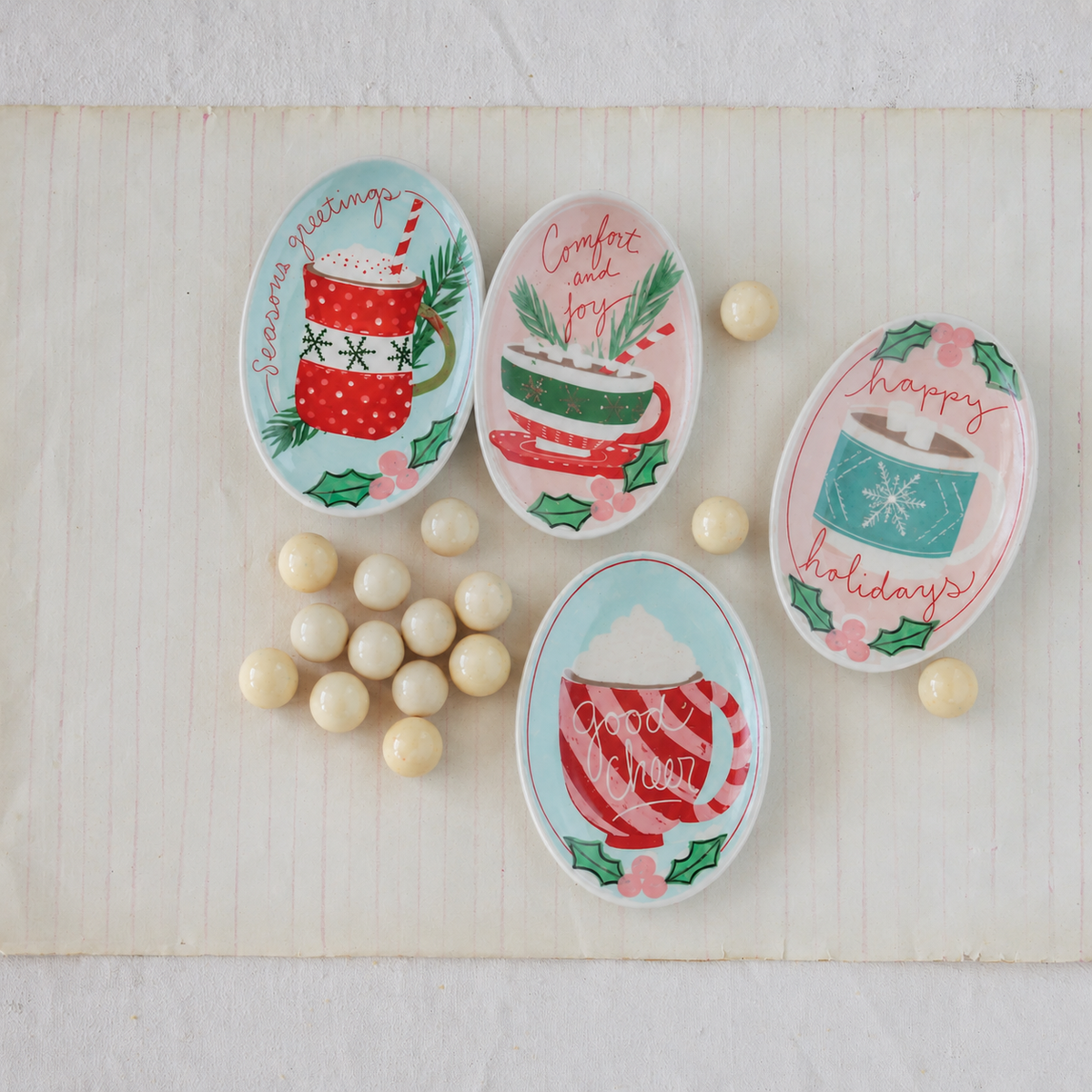 Four decorative oval ceramic dishes with holiday-themed illustrations and text, arranged on a light wood surface with small cream candies scattered around them.