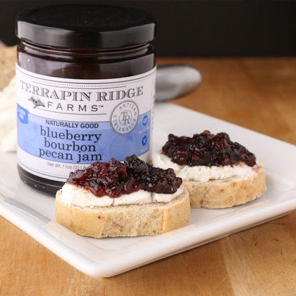 jar of Blueberry Bourbon Pecan Jam set on a tray with breads topped with cream cheese and Blueberry Bourbon Pecan Jam.