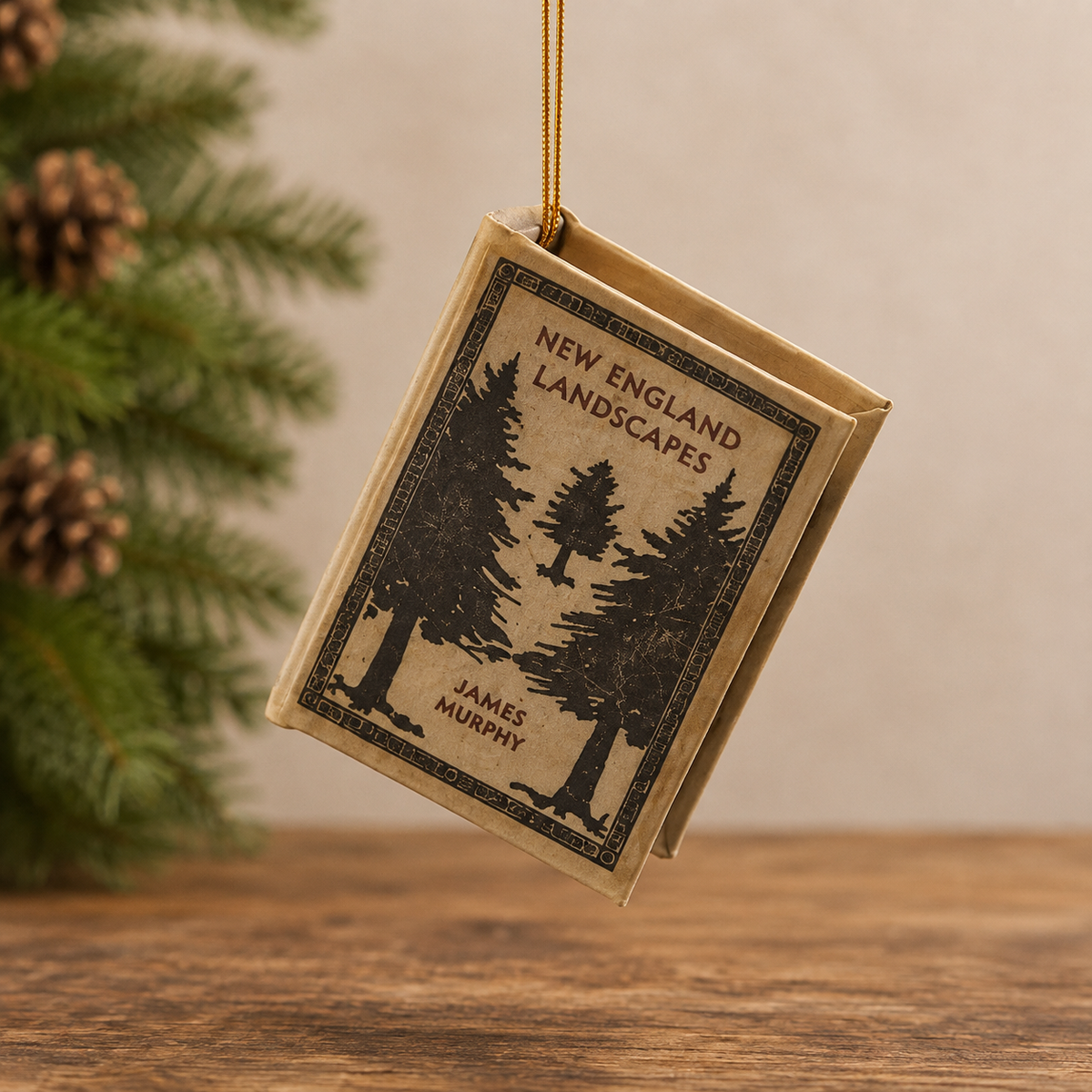 A book-shaped ornament featuring the cover art of James Murphy's 'New England Landscapes', hanging from a gold string against a warm beige background with pine branches on the left.