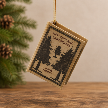 A book-shaped ornament featuring the cover art of James Murphy's 'New England Landscapes', hanging from a gold string against a warm beige background with pine branches on the left.
