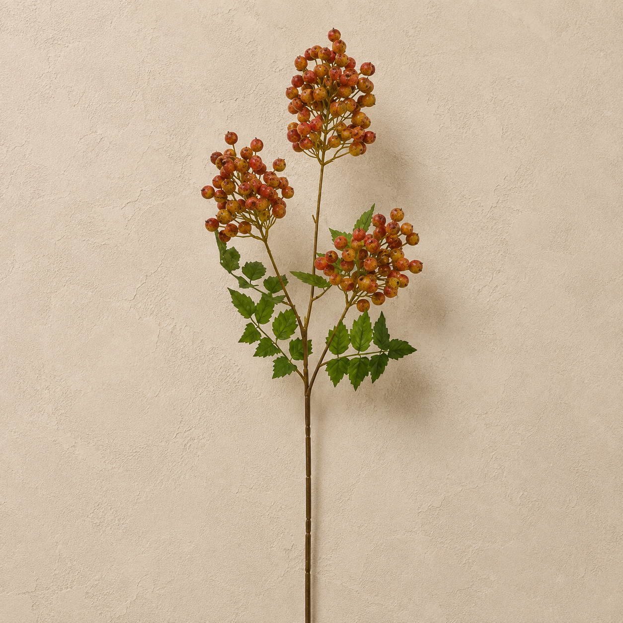A single artificial autumn stem with clusters of small orange-brown berries and green leaves, displayed against a textured beige wall.