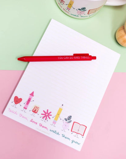 A white lined notepad with colorful cartoon crayons, flowers, and the phrase 'teach them, love them, watch them grow' at the bottom, styled on a pastel pink and mint green background with a red pen resting across the top.