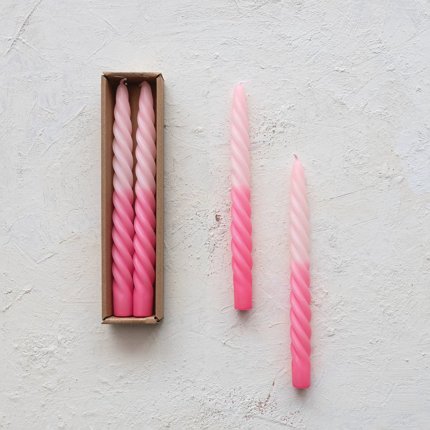 2 pink ombre twisted candles set next to a boxed set of candles on a plaster background.