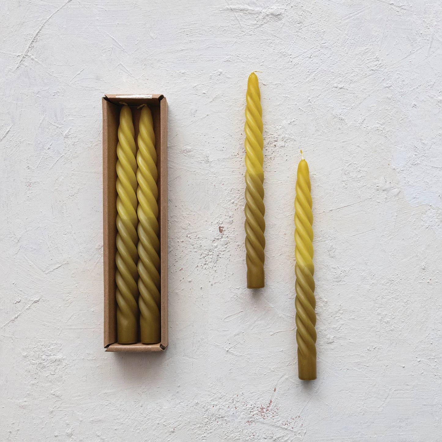  2 ombre green Twisted Taper Candles set next to a boxed set of candles on a plaster background.