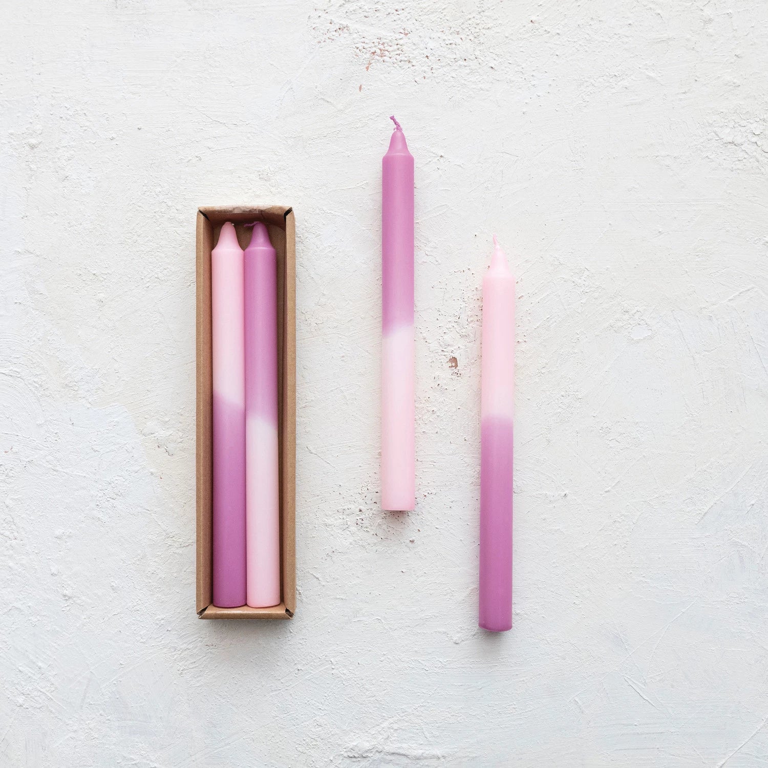 set of 2 pink and lilac taper candles set next to a boxed set of candles on a plaster background.