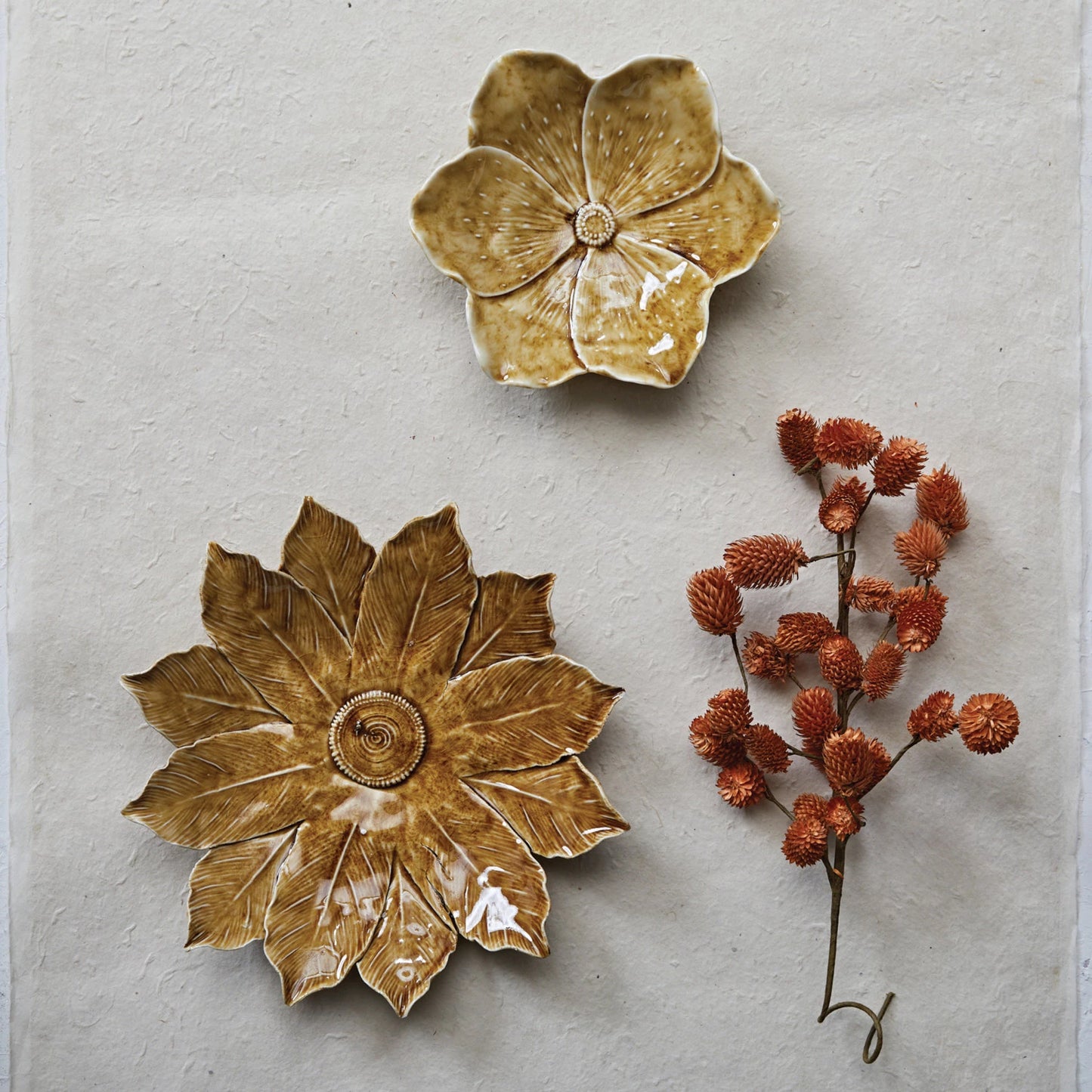both sizes of stoneware flower plates displayed next to dried orange greenery on a textured light surface