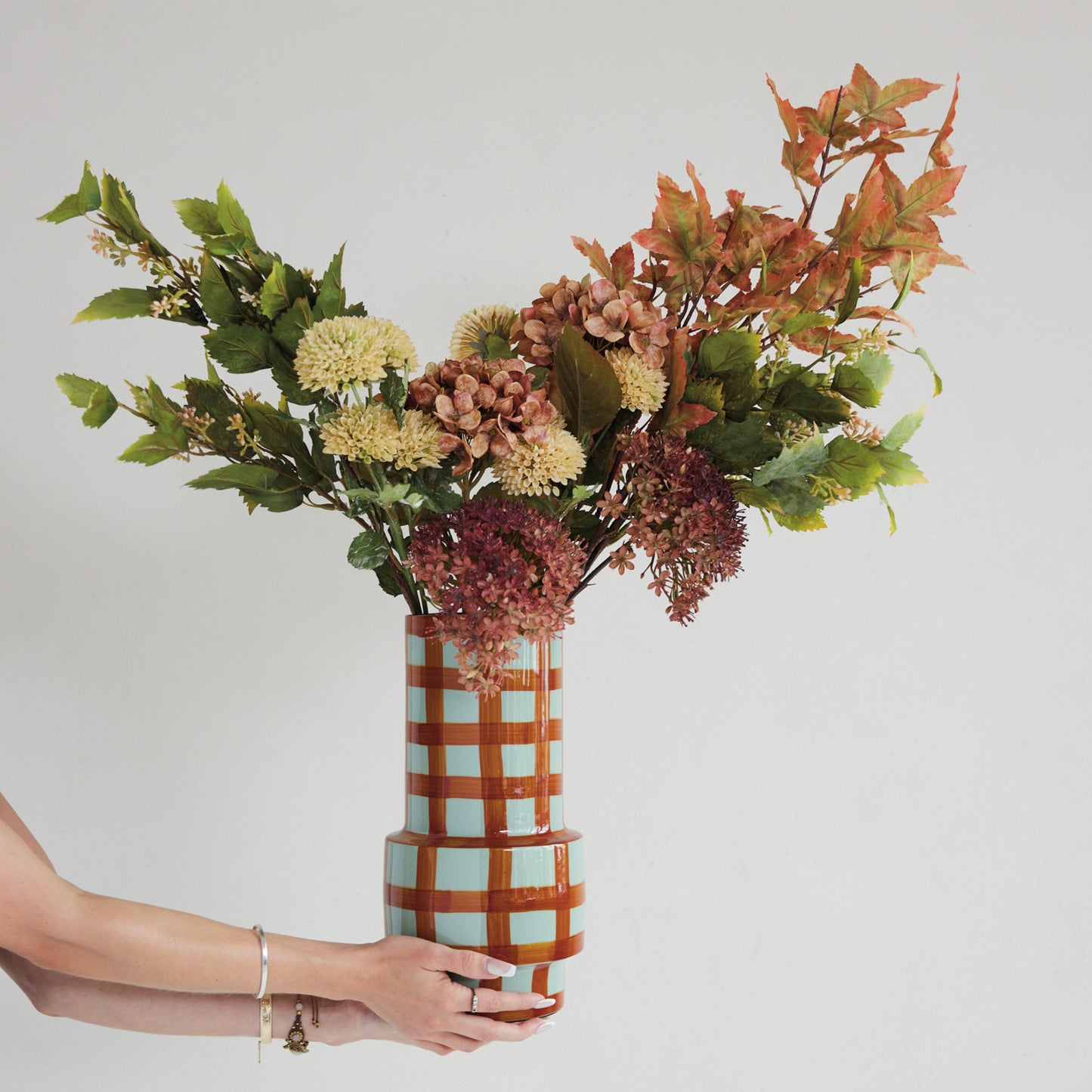 hands holding brown plaid vase filled with flowers and greenery shown on a white background