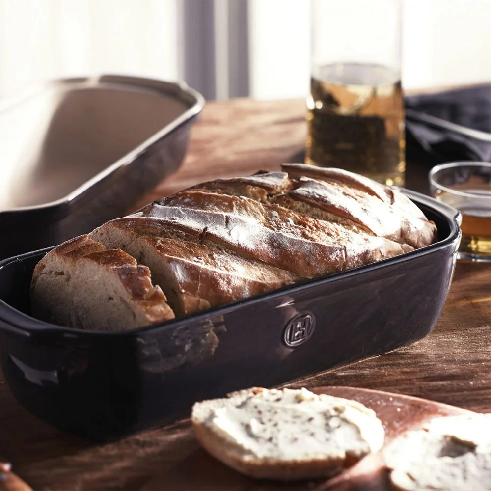 long loaf baker filled with baked bread set on a wooden table witht he lid and several slices of bread.