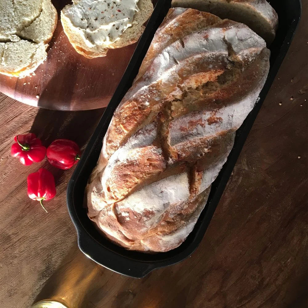 top view of long loaf baker with baked bread in it set on a wooden table with a plate of bread next to it.