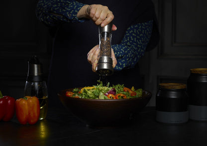 hands holding grinder and grinding pepper over a large bowl of salad.