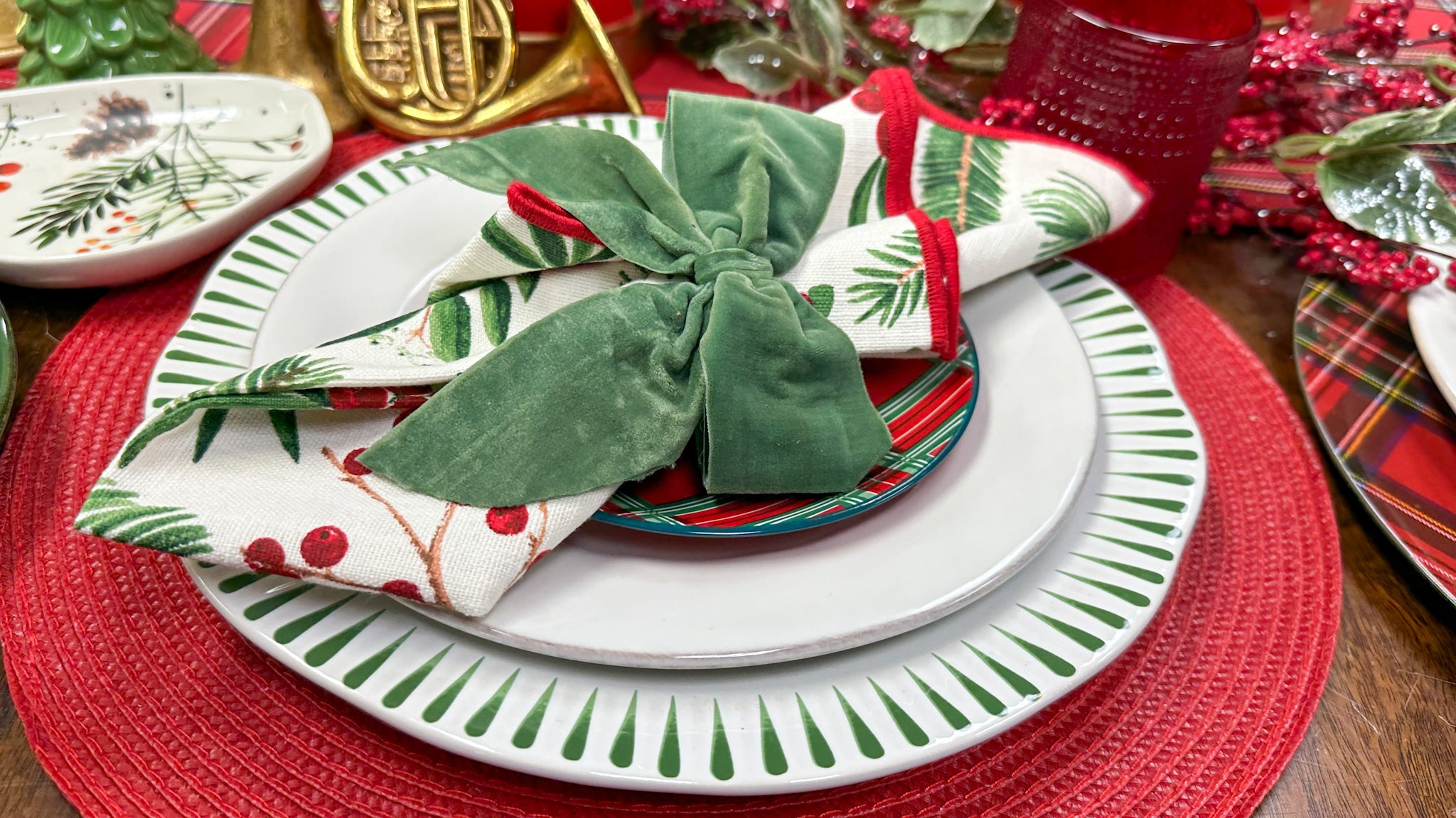 christmas themed tablescape inlcuding a stack of three holiday themed plates, a holly napkin with green velvet bow and a french horn and hobnail glass in the background.