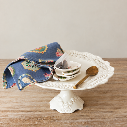 A white ceramic cake stand with a scalloped edge and floral pedestal base, styled on a wooden tabletop with a folded blue patterned cloth, a stack of small white dishes, a teabag, and a wooden spoon.