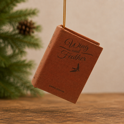 A miniature book-shaped ornament titled "Wing and Feather" by Emma Gibson, hanging from a gold string against a warm beige background with a blurred pine tree and wooden surface.