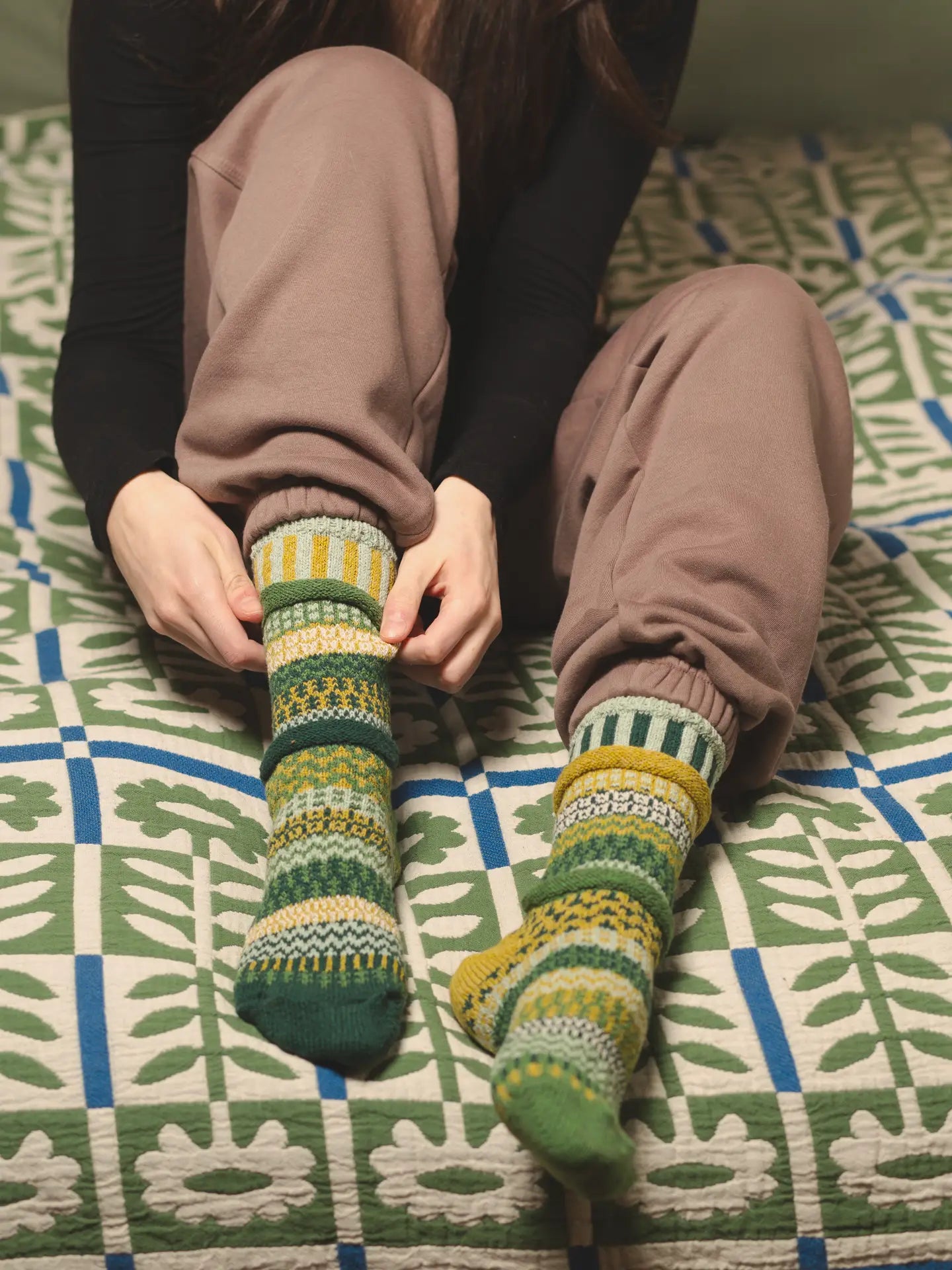 person sitting on a bed with a floral quilt on it wearing sweat pants and Fiddlehead Crew Socks