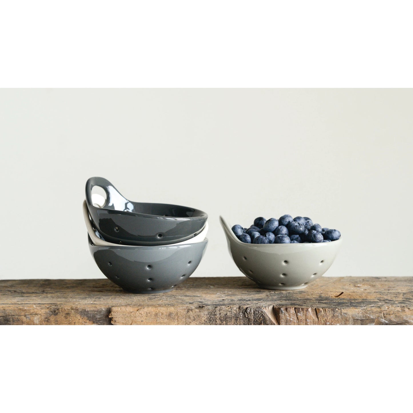 the gray stoneware berry bowl filled with blueberries and the other three colors stacked next to it and displayed on a rustic wood slab