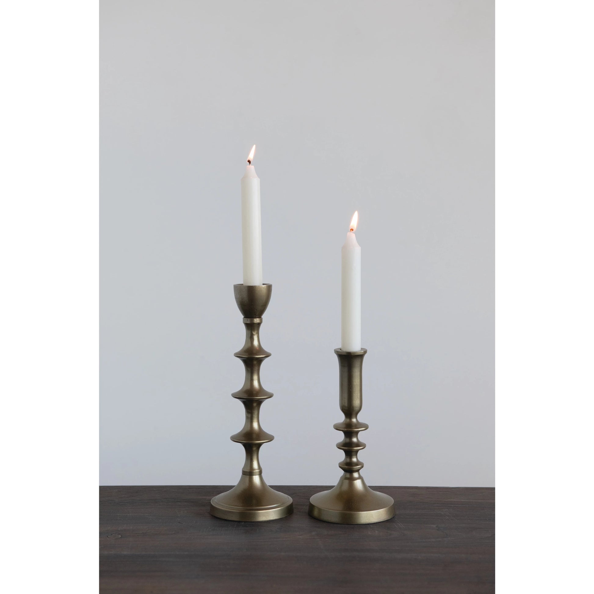 2 styles of antique gold taper holders with lit candles in them on a dark wooden table.