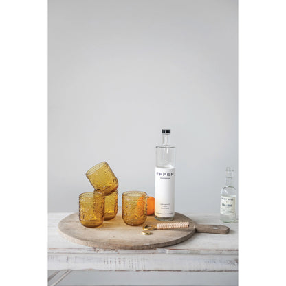 4 amber drinking glasses arranged on a wooden board with a bottle of vodka, bottle of beer, bottle opener, and an orange.