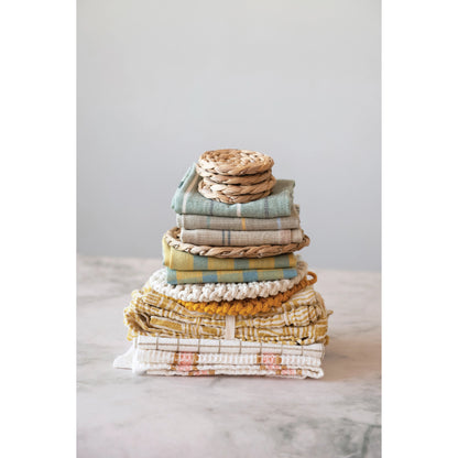 all three colors of woven cotton tea towels with stripes folded and stacked with other towels on a white and gray marble surface