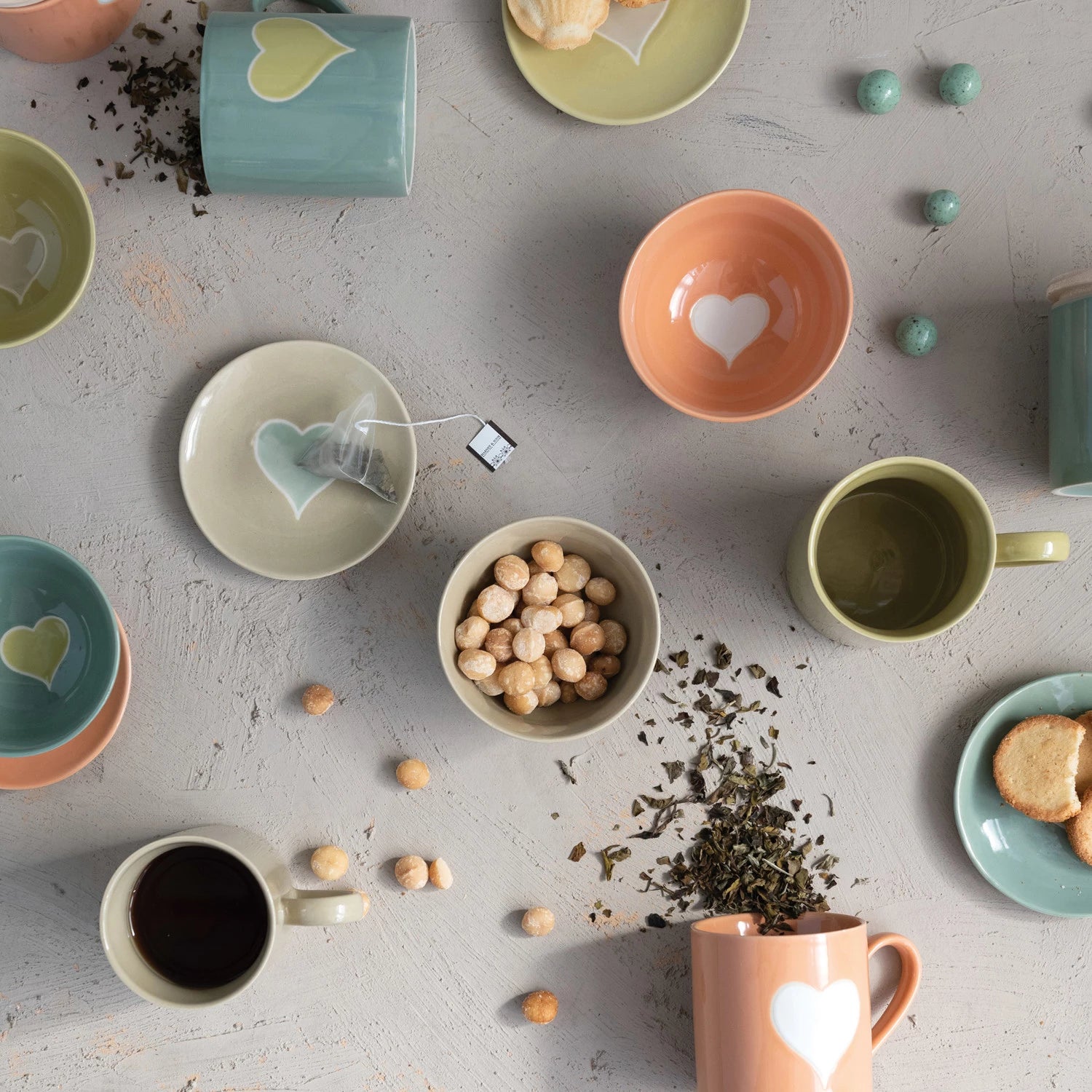 top view of all four colors of handmade stoneware mugs displayed on a table next to small bowls and small plates, tea leaves, and candy eggs